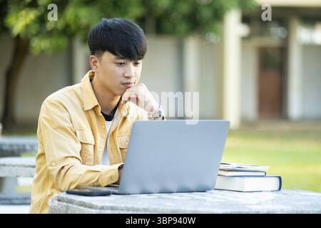A university student is absorbed in thought while studying on his laptop, sitting at an outdoor table surrounded by the tranquility of the campus Stock Photo