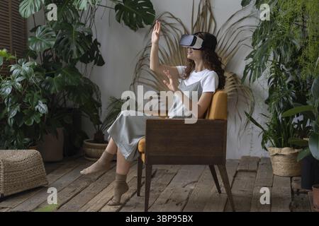 Young impressed woman wearing VR helmet sitting in armchair at greenhouse, monstera deliciosa plant on background. Hispanic female experiencing virtua Stock Photo