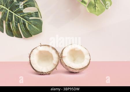 Two halves of fresh raw coconut on pink backgound with green monstera leaves on white. Trendy concept Stock Photo