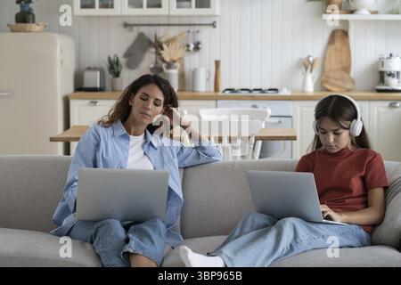 Mother freelancer doing remote work on laptop while her daughter sitting near and studying online. Modern family mom and child using digital devices f Stock Photo