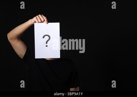 Man hiding behind paper sheet with question mark on dark background Stock Photo