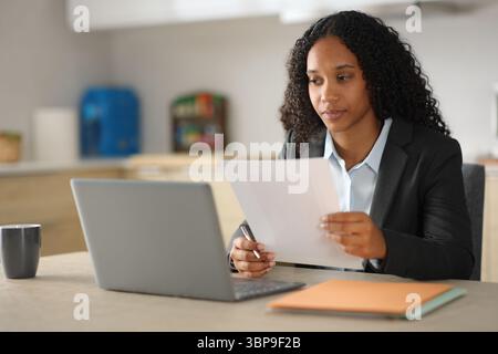 Black businesswoman checking documents and laptop in a kitchen at home Stock Photo