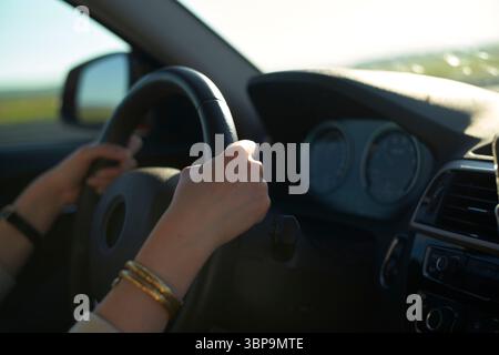 Hands firmly grip the steering wheel inside a sunlit car on an open road. Stock Photo