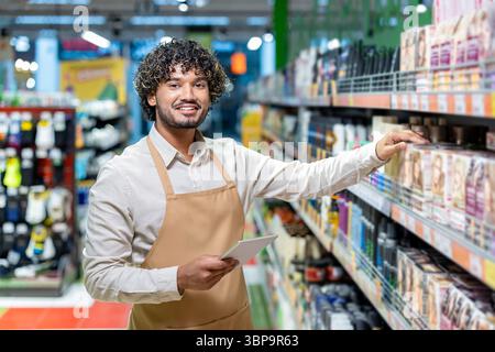 A smiling worker in a supermarket stands by shelves, checking inventory with a tablet while wearing an apron. Stock Photo
