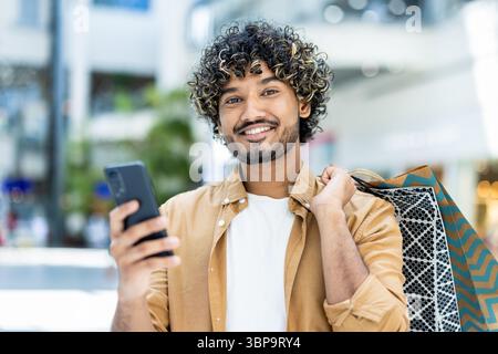 A smiling man holds his phone while carrying shopping bags, enjoying a day of shopping and using technology. Stock Photo