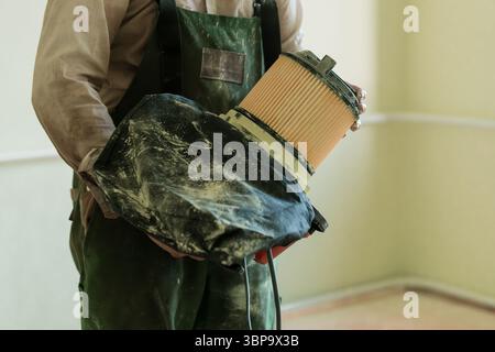 A mechanic in green overalls compares a dirty, used car air filter with a clean replacement, emphasizing vehicle maintenance, indoor workshop setting, Stock Photo