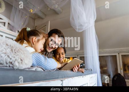 Excited single father plays with diverse, multi-ethnic daughters. Using digital tablet enjoy weekend Stock Photo