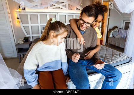 Excited single father plays with diverse, multi-ethnic daughters. Using digital tablet enjoy weekend Stock Photo