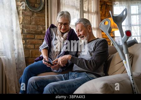 Senior wife embracing disabled husband at nursing home. Cheerful old couple using mobile phone. Stock Photo