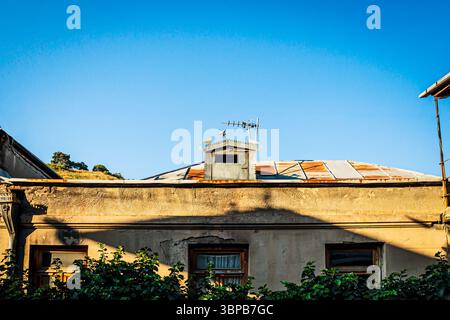 Old building with rusty metal roof and decorative rooftop ornament featuring star and birds, framed by green foliage under a bright blue sky in warm s Stock Photo