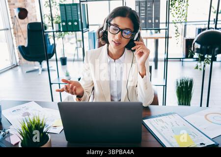 Businesswoman in a modern office talking through headphones during a video call on her laptop, multitasking with documents Stock Photo