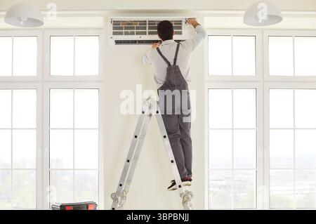 Service worker wearing overalls, standing on ladder, repairing air conditioner Stock Photo