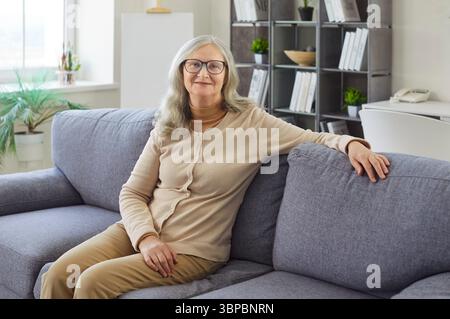 Happy senior woman sitting on sofa to enjoy cozy time in home living room, elderly lady relaxing Stock Photo
