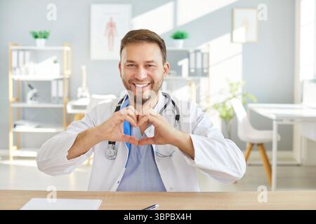 Smiling male doctor in white coat sitting at desk, making heart shape with hands in medical office. Stock Photo