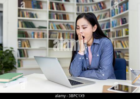 A woman yawns while working on a laptop at a desk in a library, surrounded by bookshelves. Stock Photo