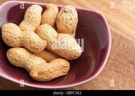 Unpeeled roasted peanuts in a red ceramic bowl on wooden brown surface. Ready to eat nut snack. Macro shot Stock Photo