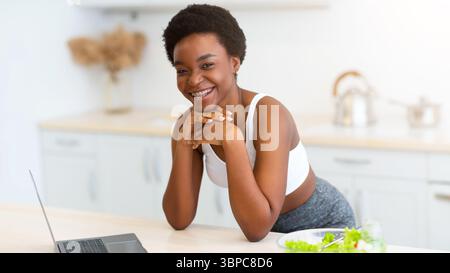 African Woman Using Laptop Browsing Recipes For Weight-Loss In Kitchen Stock Photo