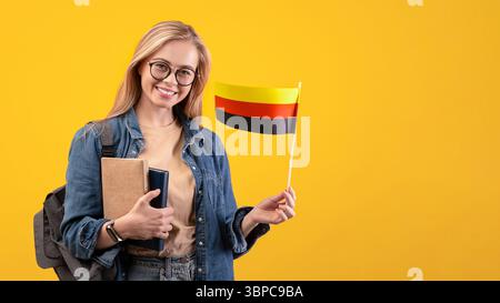 Happy young blonde lady student in casual with glasses, backpack and books holding small flag of Germany Stock Photo