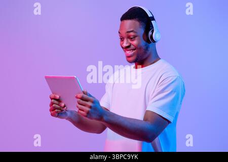 Modern Gadgets. Happy Black Man In Headset Using Digital Tablet, Neon Light Stock Photo