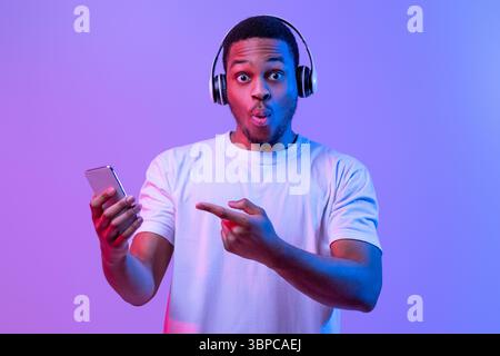 Cool App. Surprised Black Guy In Wireless Headphones Pointing At Smartphone Stock Photo