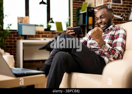 Cheerful male entrepreneur on couch, celebrating with fist raised after winning mobile game. Smiling businessman resting from remote work, holds cellphone, feeling excited from online gaming success. Stock Photo