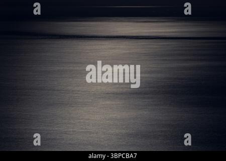 predominantly dark image of the sea at night, showing subtle ripples and a faint, diffused light on the horizon. the water has a textured, almost abst Stock Photo