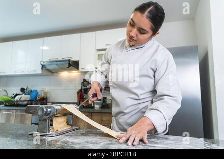 Latin woman chef using a pasta machine to make fresh pasta dough in a modern kitchen Stock Photo