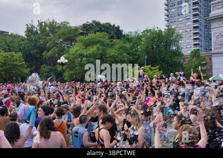 A large crowd of people, predominantly young adults, are gathered around a fountain in a park.  Many are in bathing suits or light clothing, playing i Stock Photo