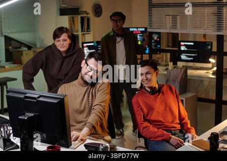 Group of people of diverse ethnicities operating computers and headsets in office at night, collaborating and monitoring screens in scam call center Stock Photo