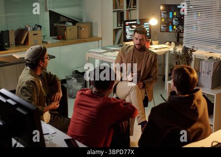 Caucasian and multiethnic individuals, sitting in office at night discussing scam operations with computer monitors displaying data in background Stock Photo