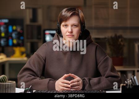 Portrait of young adult Caucasian woman wearing headset sitting at desk in office environment, looking directly at camera, working in scam call center Stock Photo