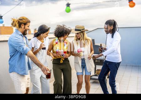 Cheerful group of diverse friends enjoying drinks, dancing, and socializing at a rooftop barbecue party Stock Photo