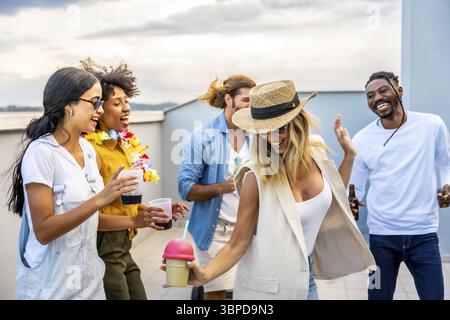 Cheerful multi ethnic friends enjoying cocktails and dancing joyfully at a vibrant rooftop party against a stunning city skyline Stock Photo