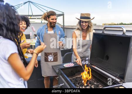 Happy friends wearing aprons are toasting marshmallows on a barbecue grill during a rooftop party Stock Photo