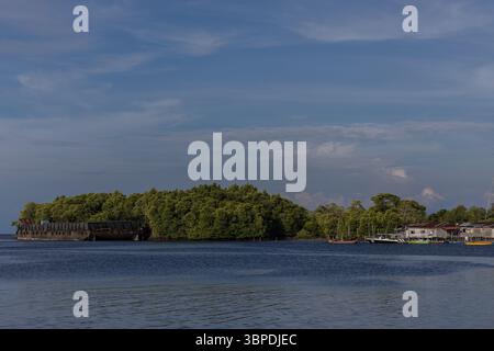 A traditional fishing boat anchored beside a wooden riverside hut surrounded by tropical mangrove trees in rural Borneo Stock Photo