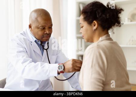 Focused black doctor man using stethoscope to examine female patients chest Stock Photo