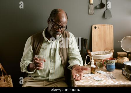 Senior Black Man Sitting at Table Taking Medication and Holding Glass of Water Stock Photo