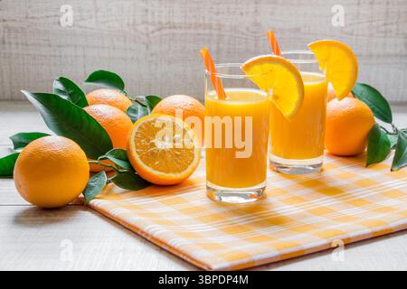 Two tall glasses of orange juice are placed next to fresh oranges on a checkered cloth. The bright kitchen setting is enhanced by natural daylight, creating a cheerful atmosphere. Stock Photo