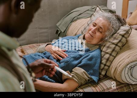 Senior Caucasian Woman Lying on Couch While Black Man Checking Temperature Stock Photo