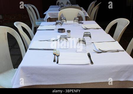 Outdoor dining table set with white tablecloth, cutlery, glassware, simple plastic chairs and a cozy atmosphereset on a sunny day. Stock Photo