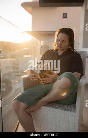 Young man with wet hair reading book on balcony chair barefoot relaxed green shorts sunset glow. Stock Photo