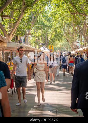 A young couple holds hands while walking with crowds of people on the famous tree-lined La Rambla promenade in Barcelona, Spain. Stock Photo