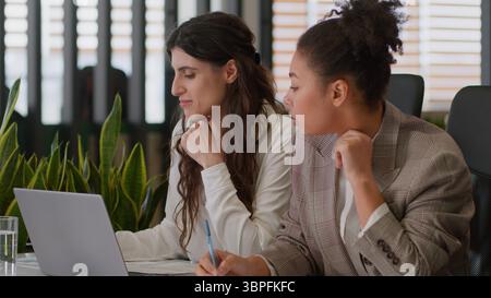 Two multiethnic women business partners female coworkers Caucasian African American colleagues ladies girls laptop work together teamwork in office Stock Photo