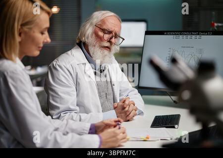 Senior Caucasian man with beard and glasses discussing research data with young adult Caucasian woman in scientific laboratory, computer monitor displaying scientific graphs Stock Photo