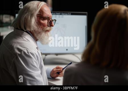 Senior Caucasian man with grey hair and beard discussing scientific data with middle aged woman in laboratory setting, both wearing lab coats, computer monitor displaying graph Stock Photo