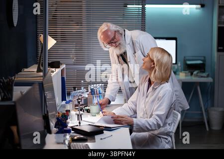 Senior Caucasian man standing and talking to middle aged Caucasian woman sitting at laboratory desk, both wearing lab coats and working with scientific equipment in laboratory setting Stock Photo
