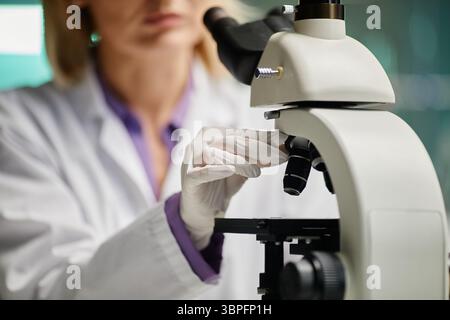 Caucasian middle aged woman adjusting microscope in scientific laboratory, wearing lab coat and gloves, focusing on specimen slide, conducting research or analysis in professional environment Stock Photo