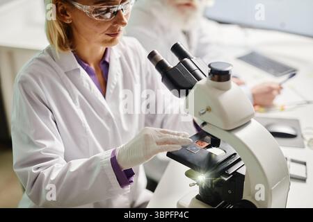 Caucasian middle aged woman examining specimen under microscope in scientific laboratory, wearing protective eyewear and gloves, older Caucasian man working in background Stock Photo