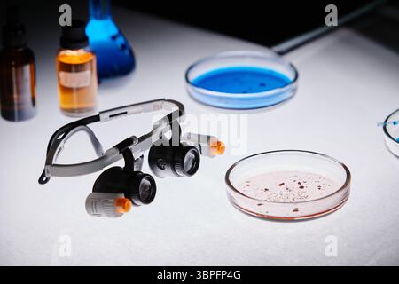 Close up of laboratory table showing protective magnifying glasses resting beside petri dishes with blue and pink liquid, scientific glassware and chemical bottles in research laboratory setting Stock Photo