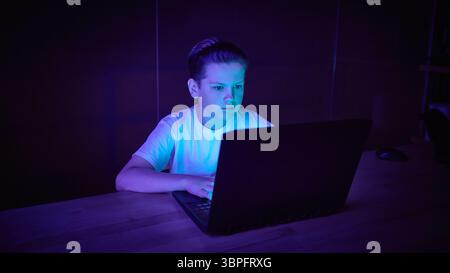 Focused boy using laptop in dark room with blue lighting Stock Photo